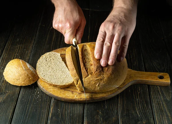 stock image The cook cuts the baked bread with a knife on the cutting board of the restaurant kitchen. The concept of healthy eating and traditional food on a black background.