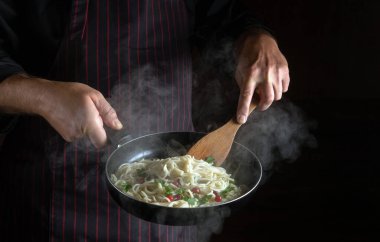 Profesyonel şef, sıcak tavada baharat ve baharatla lezzetli spagetti hazırlar. Siyah arka planda reklam için yer var. Restoran veya otel menüsü