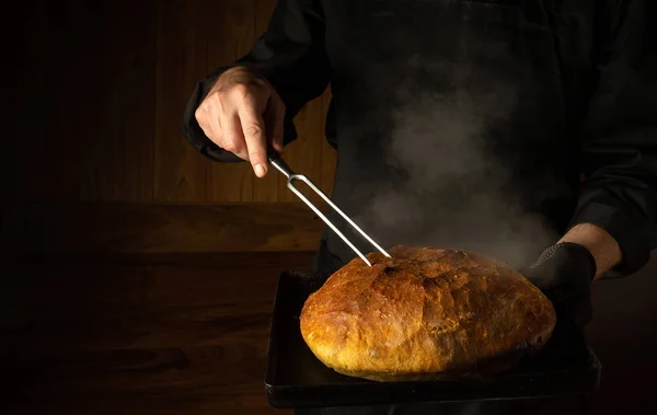 The cook bakes bread in the home bakery. Fork in the chef hand and a baking sheet with hot bread. Place for advertising on a dark background.