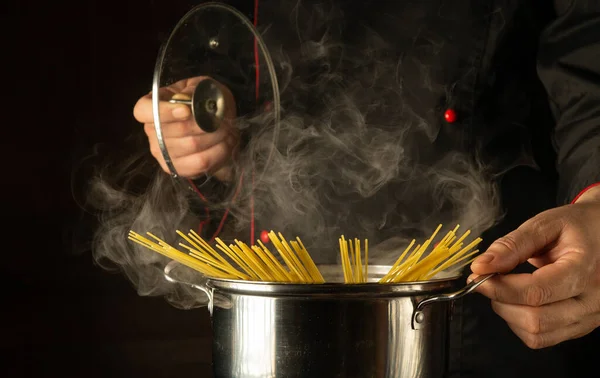 The chef cooks spaghetti in a pot with steam in the kitchen. Molecular cuisine. Free space for advertising on a dark background