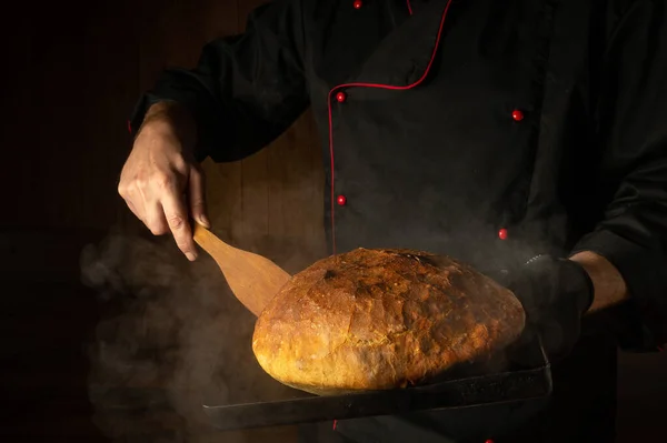 The baker bakes bread on a sheet pan in a bakery. Scraper in the chef hand. Place for menu or recipe on dark background