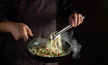 Aşçı elinde makarna olan sıcak bir tavayı tutuyor. Bir şefin elleriyle baharatlı ve baharatlı spagetti pişirmek. Siyah arkaplanda otel tarifi veya menüsü için yer