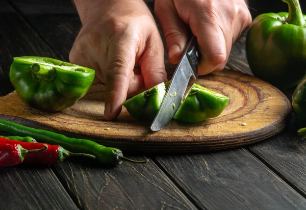 The cook cuts fresh green peppers to prepare a delicious salad at home. Close-up of a chef hands with a knife on a kitchen black table. Copy space