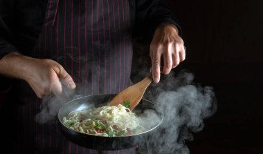 Şef, haşlanmış tavada baharatlı ve baharatlı lezzetli spagetti hazırlıyor. Karanlık arka planda reklam için yer var. Restoran veya otel menüsü