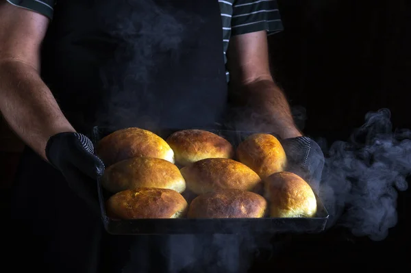 Baker holds a baking sheet with freshly baked buns or pies. Black space for menu or recipe