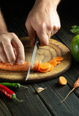 Ahşap kesme tahtasının üzerinde taze havuç doğrayan bir bıçakla şefin elinin yakın çekimi. Mutfak masasında vejetaryen yemeği pişirmek.