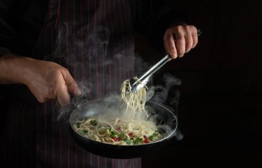 Chef cooks spaghetti in a frying pan. Recipe or menu for hotel on black background. Delicious pasta with vegetables and spices.