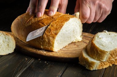 Close-up of a chef hands with a knife cutting freshly baked wheat bread before dinner. National dish on the kitchen table. Copy space