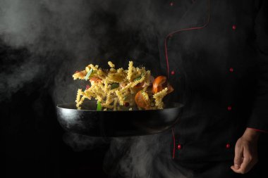 Professional chef prepares food in a hot frying pan with steam on a dark background. The concept of restaurant and hotel service. Thai food noodles with spices and vegetables.