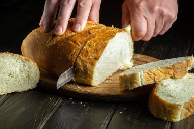 Cook's hand cut fresh bread with a knife on a kitchen cutting board. Healthy food and traditional bakery concept