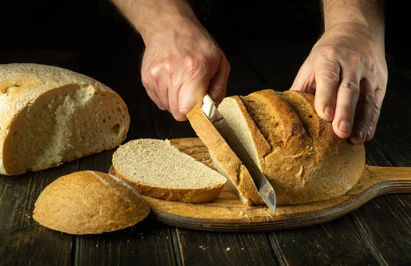 The chef cuts wheat bread on the wooden board of the bakery kitchen for tasting. Healthy food concept on black background.
