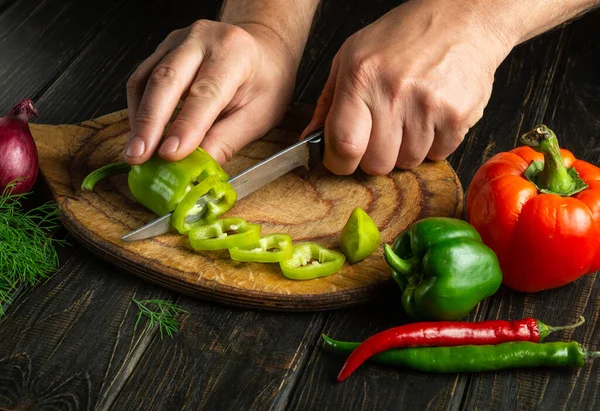 Hands of a cook with a knife cut green peppers on a cutting board to prepare a vitamin salad. Delicious peasant food. Copy space