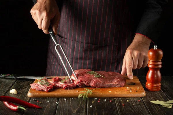 The chef with a fork in his hand prepares beef meat in the kitchen. Idea for a restaurant or hotel menu. Copy space