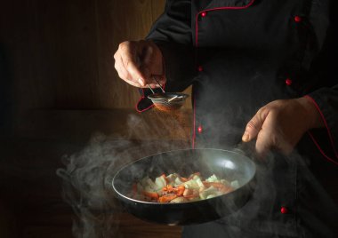 Şef kızartma tavasında kızarmış havuç ve soğana kırmızı biber ekler. Öğle yemeği için sebze pişiriyorum. Karanlık bir arkaplanda restoran ya da otel menüsü.