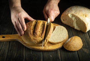 Bıçaklı bir aşçının elleri tahtanın üzerinde buğday ekmeği kesiyor. Sağlıklı yiyecekler ve arka planda geleneksel fırın konsepti. Boşluğu kopyala