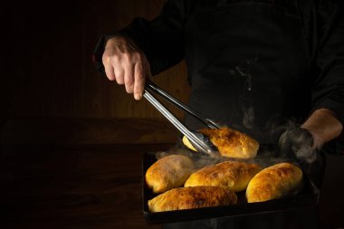 The baker bakes hot dog buns. Tongs in chef hand and baking sheet with hot pies. Place for advertising on a dark background.