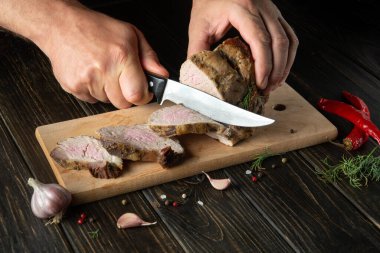 Chef slicing grilled beef meat on a cutting board. Work environment on the kitchen table with condiments or spices. Black or dark background with space for hotel menu