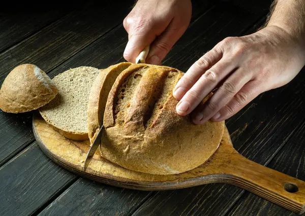 Hands of a chef with a knife cut wheat bread on a cutting wooden board. Wheat bread on the kitchen table or the healthy eating and traditional bakery concept