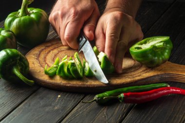 The cook slicing fresh green peppers on a wooden cutting board. Close-up of chef hands while preparing vegetarian food or adjika.