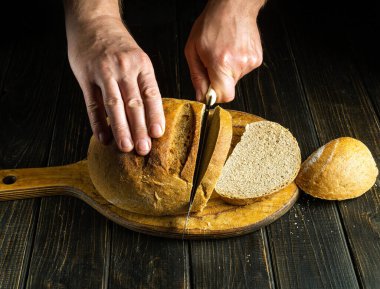  Hands of a cook with a knife cut the baked bread on the cutting board of restaurant kitchen. The concept of healthy eating and traditional food on a dark background