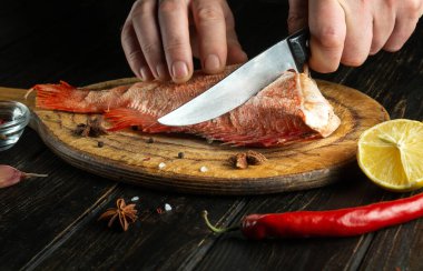 Hands of a chef cut raw Sebastes fish on the wooden cutting board of the kitchen. The concept of a delicious red fish lunch for a restaurant or hotel