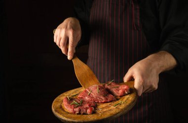 The chef turns over raw beef steaks with a wooden spatula before barbecue. Preparation for roasting meat with rosemary and spices. Black space for hotel recipe or menu