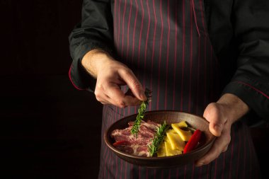 The cook puts rosemary on a plate with sliced steak and cheese. The concept of serving dishes to order by a waiter with a place for advertising on a black background.