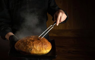 Baker bakes bread in the home bakery. Fork in the chef hand and a baking sheet with hot bread. Place for advertising on a dark background
