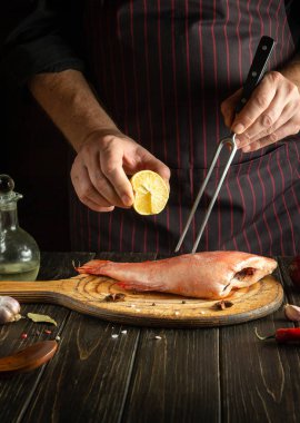 Professional chef presses lemon juice on a Sebastes fish in the kitchen before cooking. The idea of a menu or delicious recipe for a restaurant on a dark background