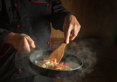 A professional chef prepares vegetables in a hot pan with steam. Space for recipe on dark background. Restaurant or hotel menu