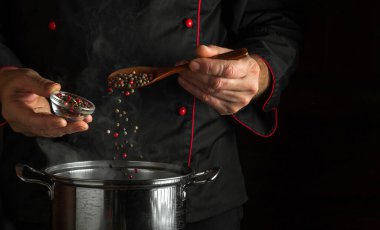 Profesyonel şef kaynayan bir tencereye güzel kokulu kuru biber ekler. Siyah arka planda reklam alanı olan bir restoran mutfağı konsepti. Aşçı elinde kaşık