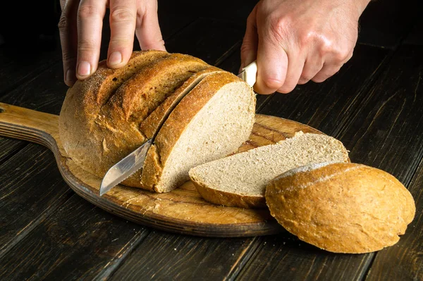 stock image The cook hands with a knife slices bread on a kitchen cutting board. The concept of healthy food in the bakery. Copy space