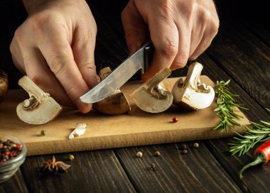 Mutfak kesim tahtasının üzerinde mantar kesen bir bıçakla bir aşçının ellerine yakın çekim. Tarif veya menü için karanlık boşluk