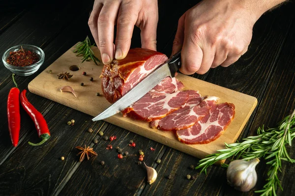 stock image Close-up of the hands of a cook with a knife cuts smoked meat or ham for lunch. The idea of fast street food