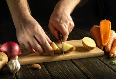 Ahşap bir kesme tahtasının üzerinde çiğ patatesle kesilmiş bir bıçakla bir aşçının ellerine yakın çekim. Restoran mutfağında sebze yemeği pişirmek..