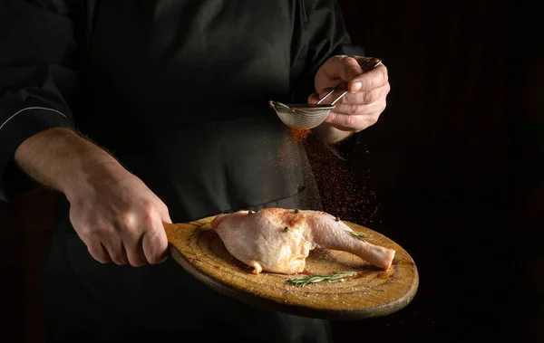Chicken leg on a cutting board in the chef\'s hand. Professional chef adds paprika. Chicken leg cooking concept on dark background