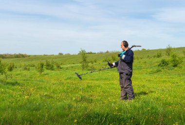 Metal detektörlü bir adam. Metal aramak için sensörlü bir adam sahada. Hazine avcısı arka plandaki mavi gökyüzünde duruyor. Hazine avcısının modern metal dedektörü var. Kürekli adam hazine arıyor..