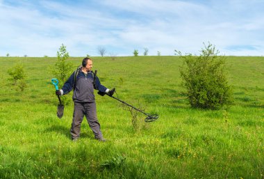 Sırtı kameraya dönük metal dedektörlü bir adam. Metal detektörlü spor kıyafetli bir adam. Yaz otlağındaki kazıcı. Yeraltında madeni para arama kavramı. Metal detektörlü kazıcı.