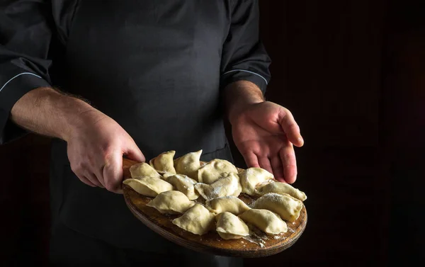 stock image Professional chef prepares dumplings in the kitchen. Cutting board with raw dumplings in the cook hands. Copy space on a dark background