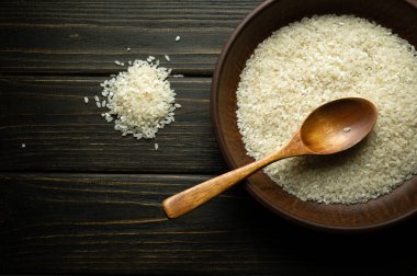 Pişirmeden önce bir tabak dolusu pirinç ve eski bir masada bir kaşık. Siyah arkaplanda tarif veya menü için yer.