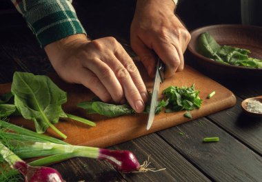 Lezzetli bir yemek hazırlamak için ıspanağı bıçakla kesen bir aşçı elinin yakın çekimi. Mutfak masasında sebzelerle çalışma ortamı