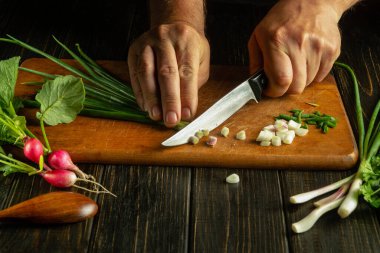 Ahşap bir kesme tahtasının üzerinde bir bıçakla taze soğan doğrayan bir şefin ellerine yakın çekim. Mutfak masasında sebzeli taze vejetaryen yemeği..