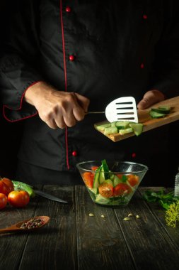Aşçı mutfakta sebze salatası hazırlıyor. Domatesli ve salatalıklı vitamin salatası pişirme kavramı. Koyu arkaplanda bir tarif için boşluk