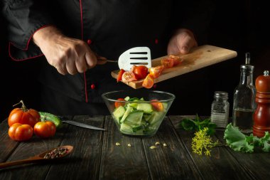 Şef mutfak masasında sebze salatası hazırlıyor. Domatesli ve salatalıklı vitamin salatası pişirme kavramı. Koyu arkaplanda bir tarif için boşluk.