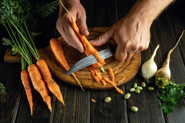The cook cleans fresh raw carrots on the kitchen table before adding them to a vegetable dish. Close-up of a chef\'s hands with a knife.