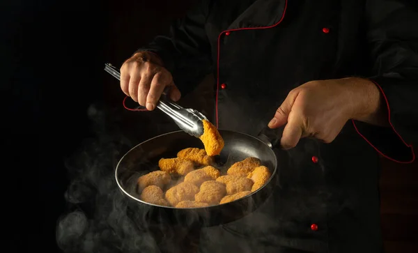 The cook prepares chicken nuggets in a frying pan. The concept of cooking nagits in the kitchen of a restaurant or hotel. Free space for advertising on a dark background.