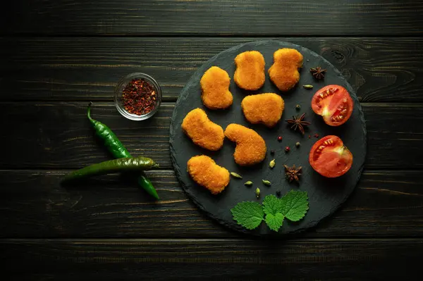 Crispy chicken nuggets on a serving round board with spices cayenne pepper and tomatoes. The concept of serving fast food on a black table in a hotel.