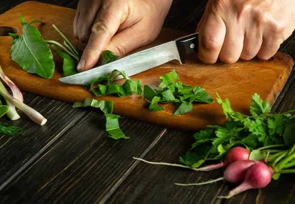 The chef uses a knife to cut sorrel on a cutting board for preparing vegetarian food. Fresh vegetables on the kitchen table.