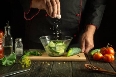 Aşçı salata için dilimlenmiş salatalığa tuz ekler. Mutfak masasında sebzelerle çalışma ortamı.