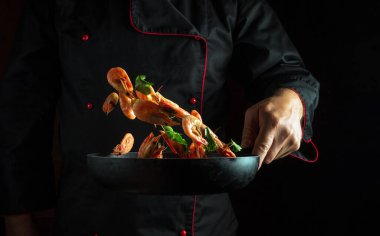 Deneyimli şef sebzeli tavada karides kızartıyor. Hareket halinde donmak ya da havada uçmak. Siyah arka planda deniz ürünleriyle yemek pişirme kavramı. Boşluğu kopyala